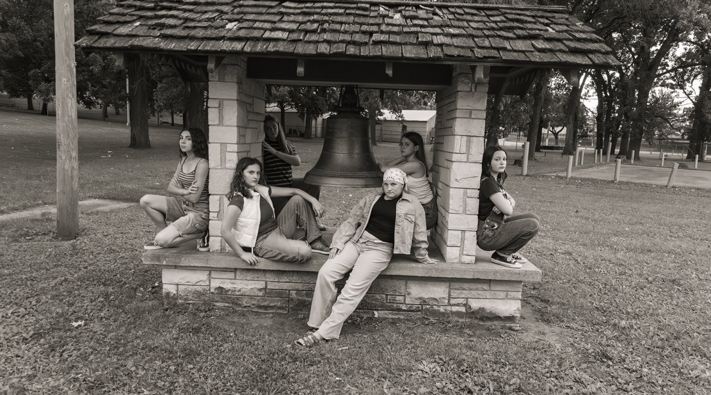 Group of young girls sit around a large bell inside a stone park gazebo, posing casually for the photo.