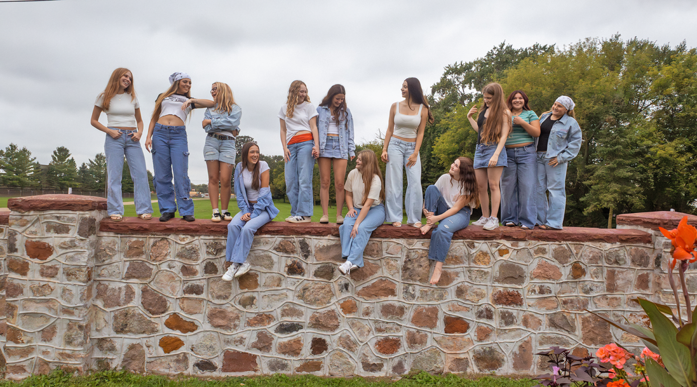 Group of teenage girls posing on a stone wall outdoors, wearing casual jeans and white tops, smiling and talking in a park setting