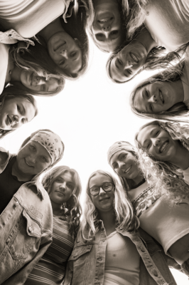 A group of friends lean in to form a circle and smile as they look down at the camera edge-to-edge around.