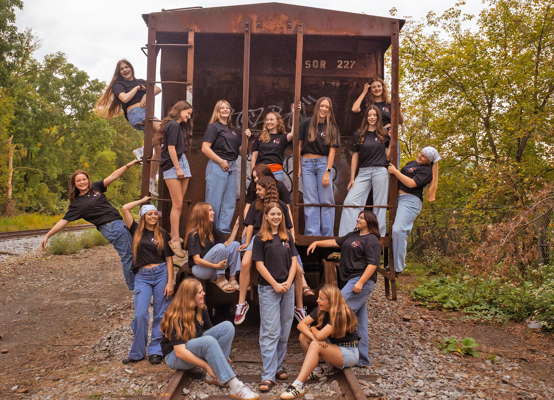 Group of young people in black shirts and jeans posing on a rusted freight car along railway tracks, smiling and looking at the camera amid trees.