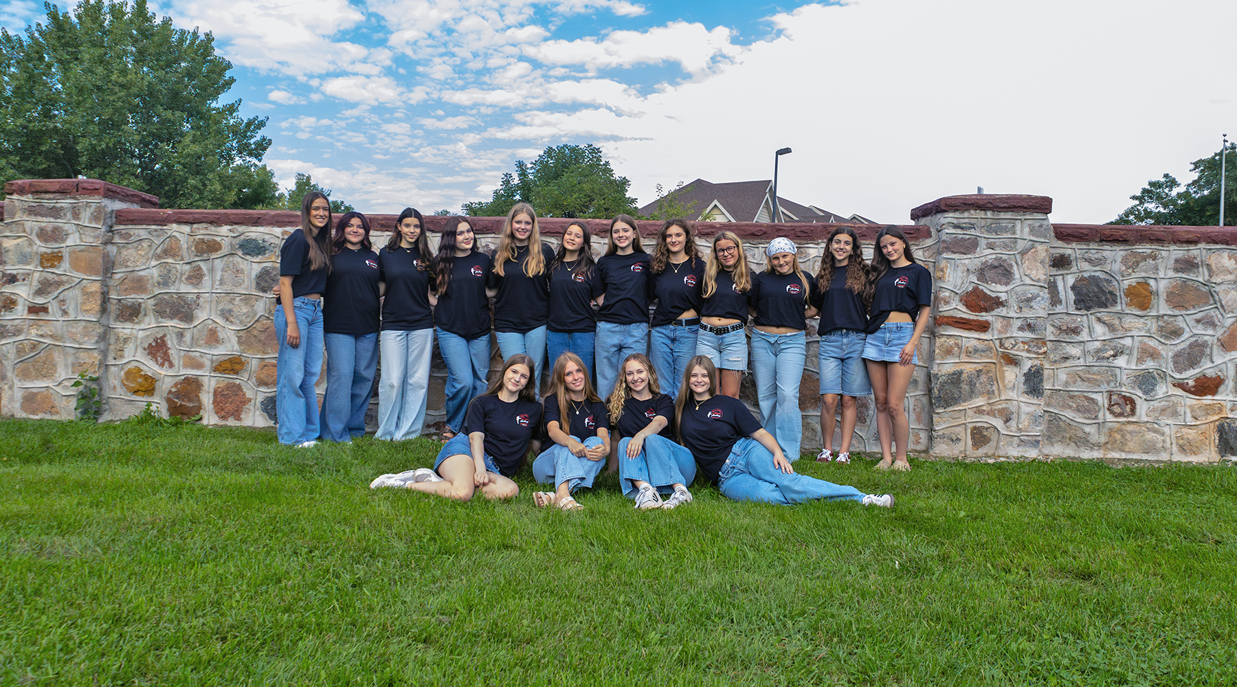 A group of girls wearing matching navy shirts and jeans posing in front of a stone wall on a sunny day, with a green lawn in the foreground.